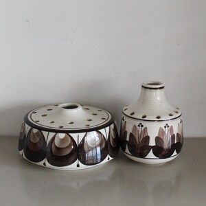 May include: Two white ceramic flower vases with brown floral designs. The vase on the left is a round, flat base with holes for flower stems. The vase on the right is a round, bulbous vase with a narrow neck.