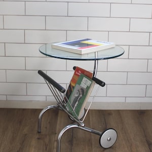 May include: A chrome-finished side table with a clear glass top and a magazine rack. The table has a single wheel and holds a book and magazines, including one with the title "History Today". The table is on a wooden floor.