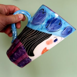 May include: A ceramic mug with a whimsical face design. The mug features a blue handle and a painted face with black hair, blue accents, and a purple and white striped pattern. The mug is held by a hand.
