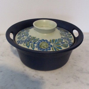 Può includere: Una casseruola in ceramica blu con un coperchio bianco che presenta un motivo floreale in tonalità di blu, verde e bianco.