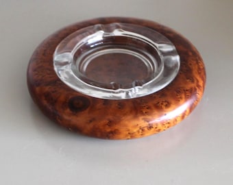 May include: Round ashtray with a clear glass center and a polished, burl wood exterior. The wood grain features a rich, warm brown color with darker, swirling patterns. The ashtray is designed for holding cigarettes or cigars.