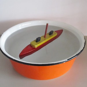 May include: A small wooden toy boat with a red hull, yellow deck, and black mast floating in a white enamel bowl with a black rim and orange exterior.