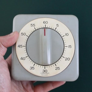 May include: A square gray kitchen timer with a circular off-white dial featuring black numbers from 5 to 60. The center has a clear rotating knob with a red indicator line. The text JUNGHANS and MADE IN GERMANY is printed at the bottom of the dial.