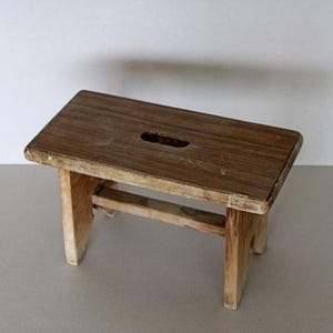 Könnte beinhalten: Ein kleiner Holzhocker mit einer rechteckigen Oberseite und einem Ausschnitt in der Mitte. Der Hocker ist aus hellbraunem Holz gefertigt und hat ein rustikales Aussehen.