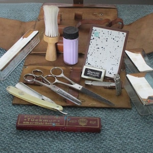 May include: A brown leather shaving kit with a variety of tools, including a razor, scissors, a brush, and a container of shaving cream. The kit also includes a small mirror and a box of razor blades. The kit is open and the tools are visible.