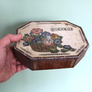 Peut inclure: Une boîte à ouvrage octogonale en bois avec un couvercle crème orné d'une illustration imprimée d'un panier tressé rempli de fleurs colorées et du mot WORKBOX. Les côtés sont en bois brun foncé avec des bordures décoratives à motifs de feuilles.
