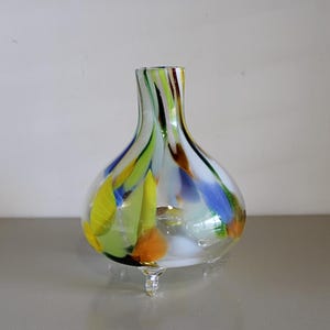 May include: A small, clear glass vase with a round body and a narrow neck. The vase has a colorful, abstract design with streaks of yellow, green, blue, and orange glass. The vase sits on three small, clear glass feet.