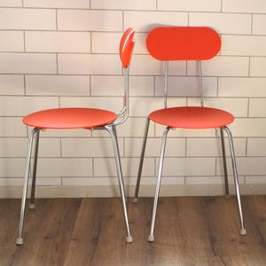 Könnte beinhalten: Zwei Stühle im Retro-Stil mit leuchtend orangefarbenen Sitzflächen und Rückenlehnen. Die Stühle haben schlanke, chromfarbene Beine und eine geschwungene Rückenlehne. Die Stühle stehen vor einer weißen Backsteinmauer und einem Holzboden.