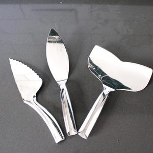 Puede incluir: Tres utensilios de servicio brillantes de acero inoxidable sobre una superficie gris oscuro. El juego incluye una paleta para tartas con borde dentado, un cuchillo para pasteles y una cuchara para servir, todos con mangos curvos.