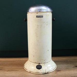Può includere: Un'antica pattumiera in metallo bianco sporco con coperchio argentato. Il bidone cilindrico ha un pedale nero e due maniglie nere. Il bidone ha una piccola etichetta rettangolare in alto. Su un pavimento in legno.