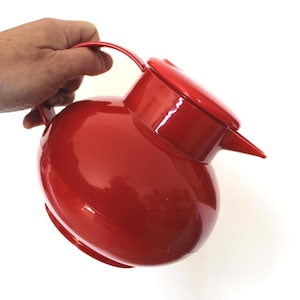 May include: A vibrant red pitcher with a rounded form, a handle, and a spout. The pitcher is held by a hand, indicating its scale and purpose. The design is straightforward and practical, ideal for serving drinks.