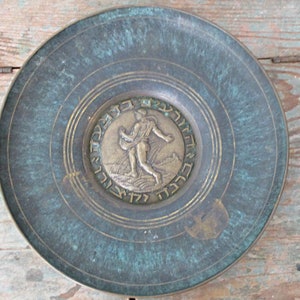 May include: A round, antique bronze plate with a central medallion featuring a figure and Hebrew text. The plate has a verdigris patina and concentric circle detailing. The plate's design suggests a historical or religious theme.