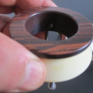 May include: A brown and white wooden ring with a metal screw in the center. The ring is made of two pieces, a top piece with a wood grain pattern and a bottom piece that is white.