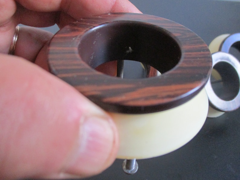 May include: A brown and white wooden ring with a metal screw in the center. The ring is made of two pieces, a top piece with a wood grain pattern and a bottom piece that is white.
