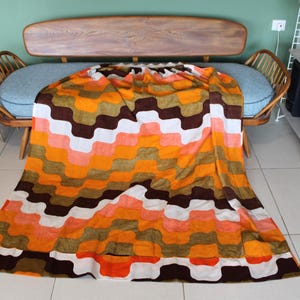May include: A colorful, patterned blanket with a retro design. The blanket features a repeating wave pattern in shades of orange, brown, white, and peach. The blanket is draped over a wooden and blue upholstered sofa, creating a vintage aesthetic.