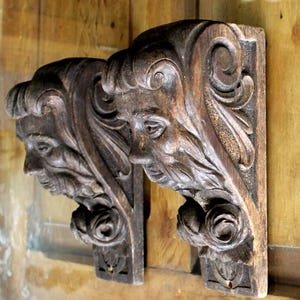 May include: A pair of dark brown wooden corbels featuring detailed carvings of human faces with flowing hair and scrollwork patterns. Each architectural bracket displays a sculpted visage in profile, integrated into a decorative support structure.