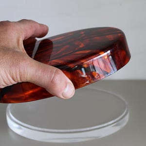 May include: A round, brown and orange, tortoise shell-patterned lid for a decorative box or container. The lid is made of a glossy, hard plastic and is being held by a hand.