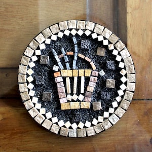Pode incluir: Uma base de mosaico redonda com um design de cesta de flores estilizada. A base tem um fundo cinza escuro com uma cesta de mosaico em tons de bege, marrom e amarelo. A borda é feita de pequenas telhas quadradas em bege e branco.