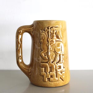 Può includere: Una tazza in ceramica marrone con manico e un design in rilievo dettagliato. Il design presenta una figura a cavallo con una lancia, con testo sottostante. La tazza ha una finitura lucida.