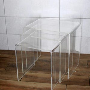 May include: Set of three clear acrylic nesting tables. Each table has a square top and a rectangular base. The tables are designed to fit inside each other, offering a space-saving solution. The tables are made of transparent acrylic.