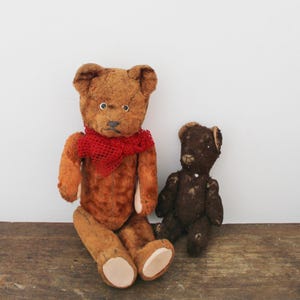 May include: Two vintage teddy bears. The larger bear is brown with a red scarf and cream-coloured paw pads. The smaller bear is dark brown. Both bears have button eyes and stitched features. They are sitting on a wooden surface.