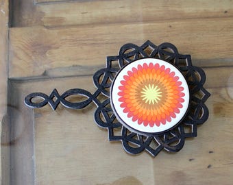 May include: A decorative trivet with a black, geometric design and a circular ceramic insert. The insert features a vibrant floral pattern in shades of yellow, orange, and red. The trivet has a handle for easy use.
