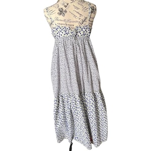 Puede incluir: Un vestido midi sin mangas con base blanca y un pequeño estampado floral azul. El corpiño tiene un diseño fruncido con tirantes finos, mientras que la falda incluye un dobladillo escalonado, creando una silueta fluida en la prenda.