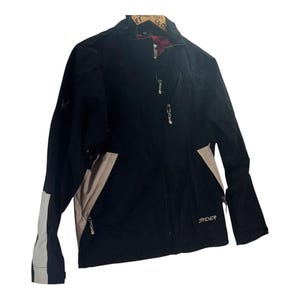 Puede incluir: Chaqueta de esquí Spyder negra con cremallera completa, paneles beige y detalles blancos en las mangas. La chaqueta tiene un cuello alto y múltiples bolsillos con cremallera. El logotipo de Spyder es visible.