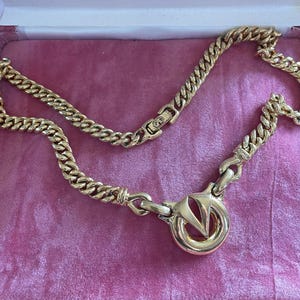May include: A gold-toned chain necklace with a large circular pendant featuring a stylized "V" design. The necklace has a thick, curb chain style and is displayed on a pink velvet surface.