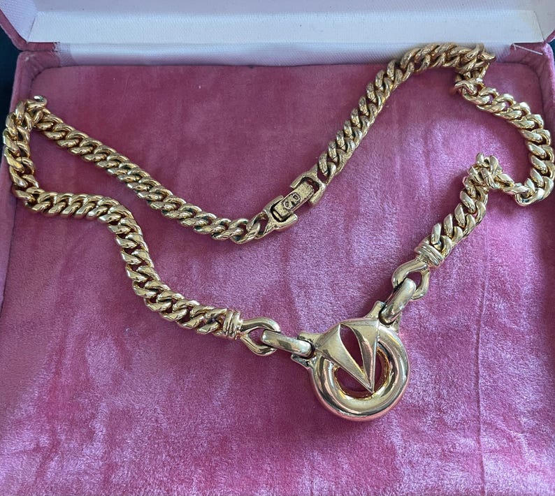 May include: A gold-toned chain necklace with a large circular pendant featuring a stylized "V" design. The necklace has a thick, curb chain style and is displayed on a pink velvet surface.