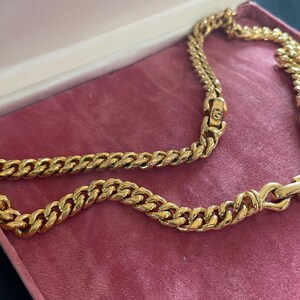 May include: A gold-toned chain necklace with a curb link design, resting inside a pink jewelry box. The necklace is made of multiple interlocking links, creating a bold and eye-catching accessory. The box has a soft, velvet-like texture.