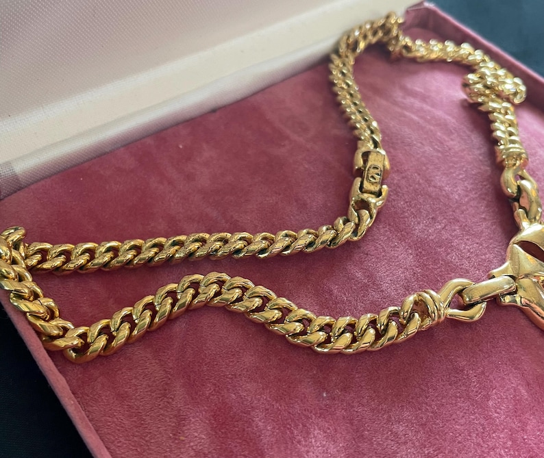May include: A gold-toned chain necklace with a curb link design, resting inside a pink jewelry box. The necklace is made of multiple interlocking links, creating a bold and eye-catching accessory. The box has a soft, velvet-like texture.