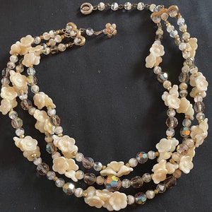 May include: A three-strand necklace featuring white flower-shaped beads and clear and brown iridescent beads.