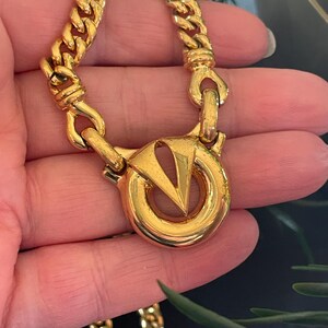 May include: A gold-toned chain necklace with a large, circular clasp featuring a stylized "V" design. The chain has a thick, curb link style. The necklace is shown against a neutral background, highlighting its metallic sheen.