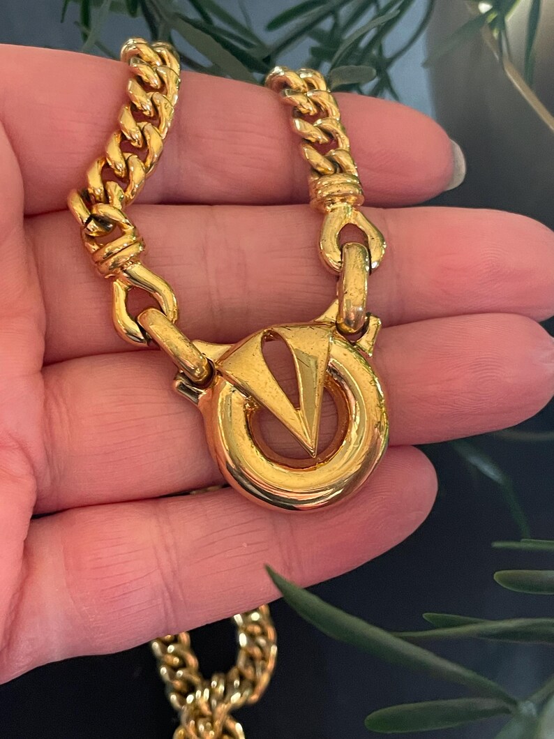 May include: A gold-toned chain necklace with a large, circular clasp featuring a stylized "V" design. The chain has a thick, curb link style. The necklace is shown against a neutral background, highlighting its metallic sheen.