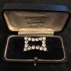 May include: A rectangular brooch with clear, sparkling stones set in a silver-toned metal frame. The brooch is displayed inside an open, vintage black jewelry box with a blue velvet interior. The box has text that reads "Fattorini & Sons Limited, Goldsmiths, Bradford."