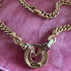 May include: A gold-toned necklace with a thick chain design. The pendant features a circular design with the brand name "VALENTINO" inscribed. The necklace is displayed on a pink surface.