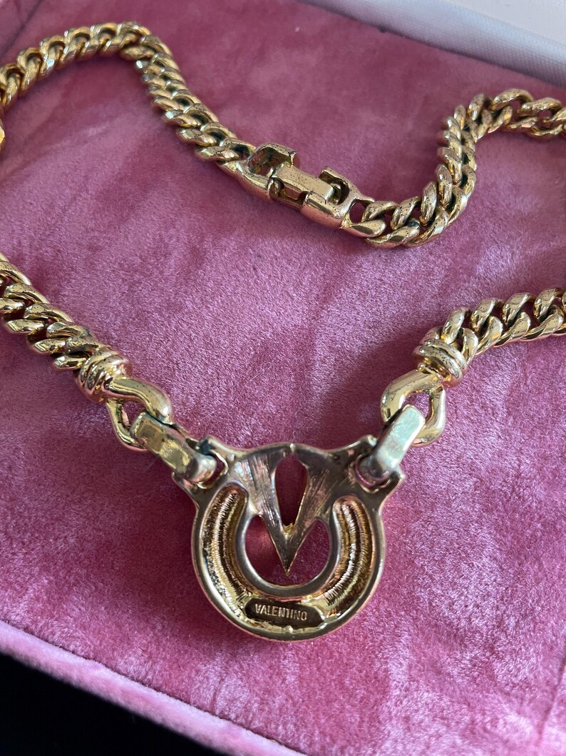May include: A gold-toned necklace with a thick chain design. The pendant features a circular design with the brand name "VALENTINO" inscribed. The necklace is displayed on a pink surface.