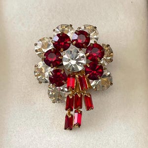 Pode incluir: Broche vintage com um design floral. O broche apresenta um aglomerado de strass transparentes e vermelho rubi, com uma pedra central transparente. Detalhes em metal dourado e pedras retangulares vermelhas completam o design.