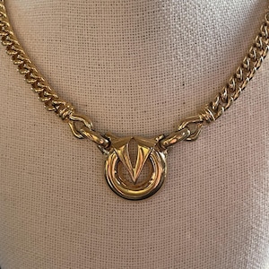 May include: Gold-tone necklace with a chunky chain and a circular pendant. The pendant features a stylized V-shaped design within a ring. The necklace is displayed on a neutral-colored surface.