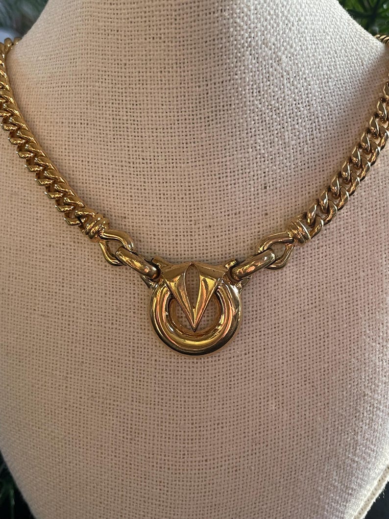 May include: Gold-tone necklace with a chunky chain and a circular pendant. The pendant features a stylized V-shaped design within a ring. The necklace is displayed on a neutral-colored surface.