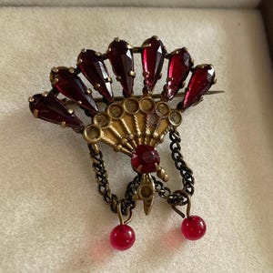 Peut inclure: Broche vintage en forme d'éventail, avec un cadre en métal doré et des accents en verre rouge foncé. La broche présente une pierre rouge centrale, des chaînes pendantes et deux perles rouges. Un accessoire rétro.