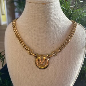 May include: A gold-toned necklace with a prominent circular pendant featuring a stylized V-shaped design. The necklace has a curb chain style and is displayed on a mannequin. The jewelry is a vintage piece.
