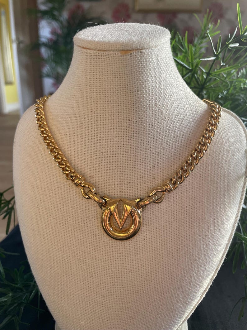 May include: A gold-toned necklace with a prominent circular pendant featuring a stylized V-shaped design. The necklace has a curb chain style and is displayed on a mannequin. The jewelry is a vintage piece.