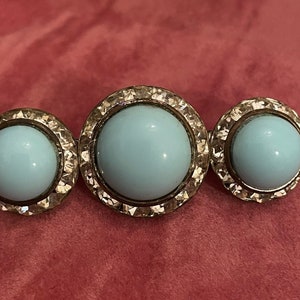 May include: Three round, silver-toned brooches with a light blue cabochon center. Each brooch has a decorative, textured border.