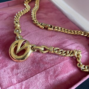 May include: A gold-toned necklace with a large pendant featuring a stylized "V" within a circle. The chain is a curb link design, and the necklace rests in a pink velvet jewelry box. The clasp is visible.
