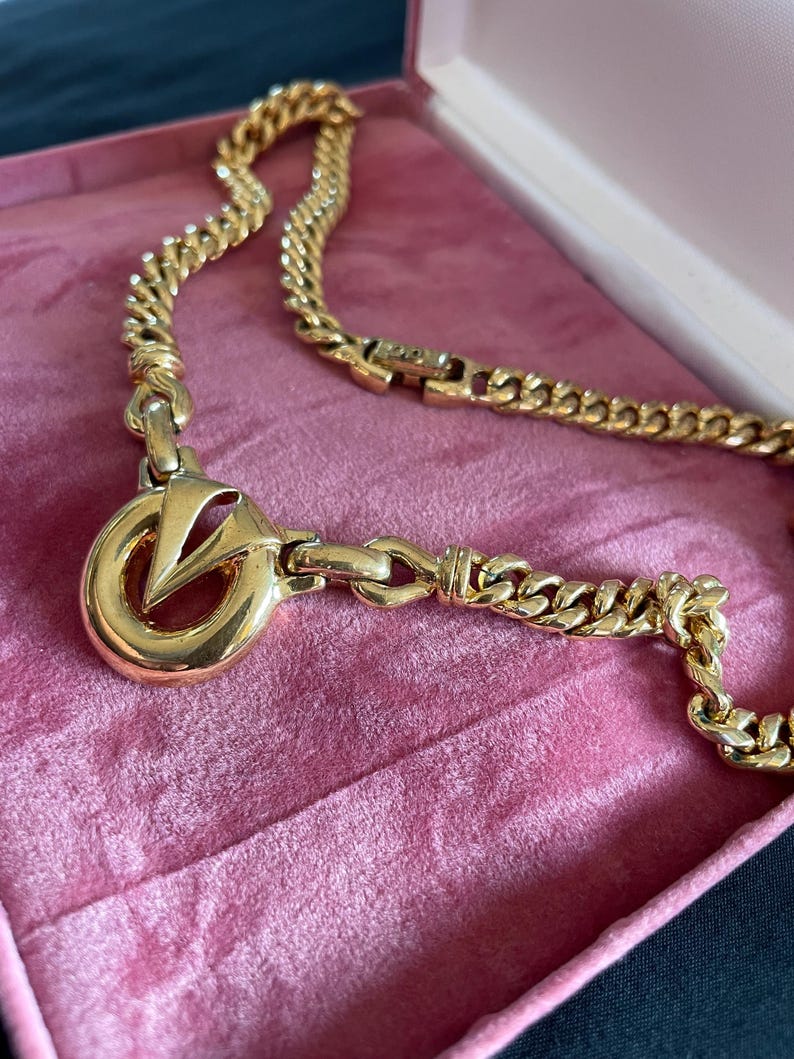 May include: A gold-toned necklace with a large pendant featuring a stylized "V" within a circle. The chain is a curb link design, and the necklace rests in a pink velvet jewelry box. The clasp is visible.