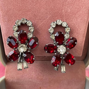 Może przedstawiać: Para zabytkowych kolczyków na klips. Każdy kolczyk ma kwiatowy wzór z owalnymi kamieniami w kolorze rubinu i przezroczystymi okrągłymi i prostokątnymi kryształami. Kolczyki są osadzone w metalu w kolorze srebrnym.
