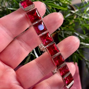 May include: A silver bracelet with a row of square-shaped red gemstones.