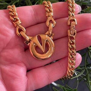 May include: A gold-toned necklace and bracelet set. The necklace features a large circular pendant with a V-shaped design. The chain links are thick and rectangular. The bracelet has a matching chain and clasp. The jewelry is displayed against a green background.
